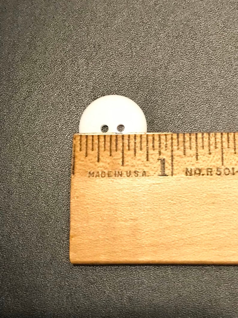 May include: A white plastic button with two holes, measuring approximately 1 inch in diameter. The button is placed next to a ruler for size reference.