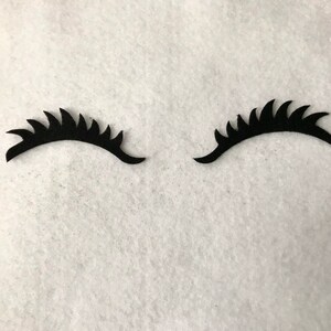 May include: Two black felt eyelashes with a jagged edge, on a white background.