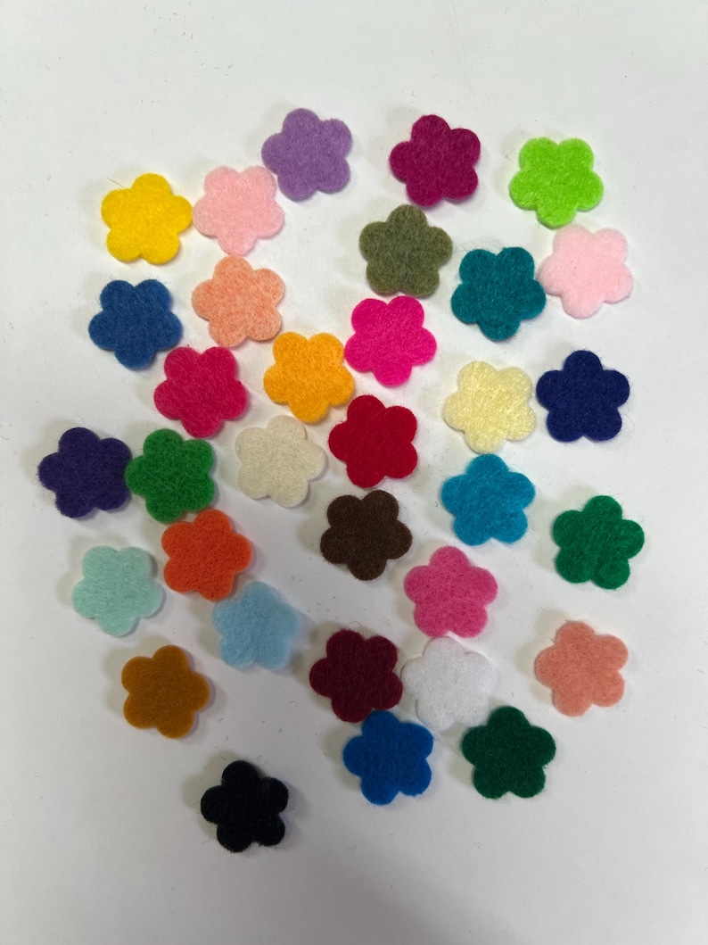 May include: A collection of small, five-petaled flower shapes made of felt in a wide variety of vibrant colors including red, blue, yellow, green, orange, purple, pink, brown, black, white, and various pastel shades scattered across the frame.