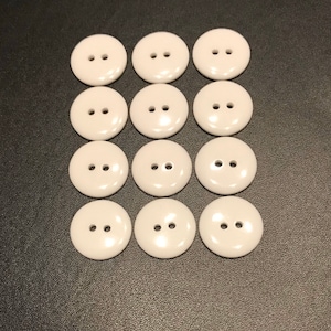 May include: Twelve white plastic buttons with two holes each. The buttons are arranged in three rows of four buttons.