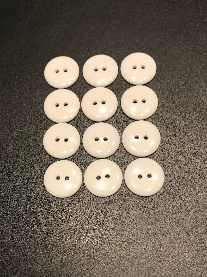 May include: Twelve white plastic buttons with two holes each. The buttons are arranged in three rows of four buttons.