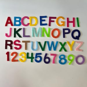 May include: Colorful felt alphabet letters and numbers arranged on a white surface. The letters are in various colors, including red, blue, yellow, and green. Numbers 1 through 0 are also displayed in a variety of colors.