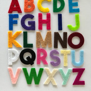 May include: A collection of colorful felt alphabet letters arranged on a white surface. The letters are in various bright colors, including red, orange, yellow, green, blue, purple, pink, brown, and white. The letters are capital letters.