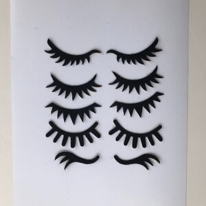 May include: Eight black paper cutouts of eyelashes in various styles, including closed eyes, open eyes, and single eyelashes. The cutouts are arranged in two rows of four on a white background.