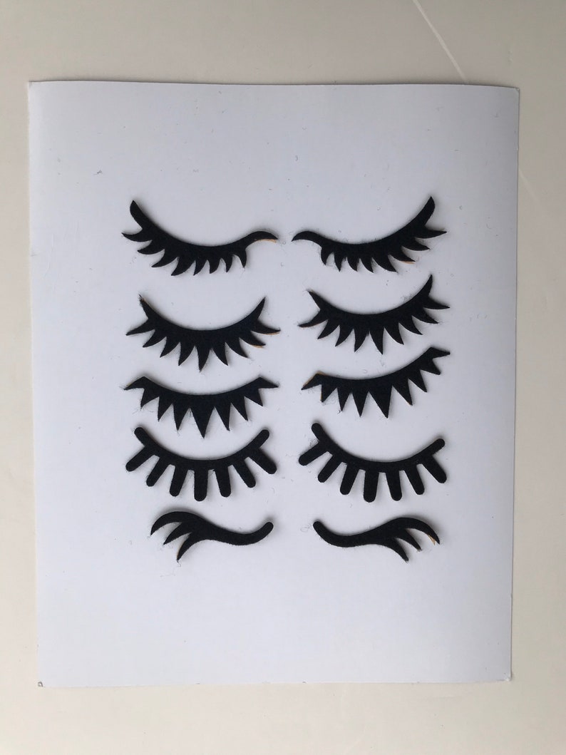 May include: Eight black paper cutouts of eyelashes in various styles, including closed eyes, open eyes, and single eyelashes. The cutouts are arranged in two rows of four on a white background.