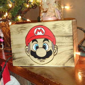 May include: A wooden box with a painted image of Super Mario on the front. The box is sitting on a brick fireplace mantel with Christmas lights and stockings hanging from it.