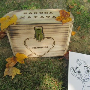 Peut inclure: Une boîte en bois avec les mots "Hakuna Matata" et "Souvenirs" gravés dessus. La boîte est décorée d'un cœur et d'un petit trou de serrure en métal. La boîte est posée sur un lit d'herbe verte et de feuilles.