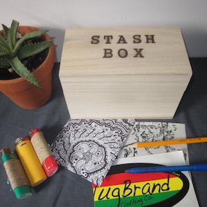 Op de afbeelding: Een houten doos met de woorden "STASH BOX" gegraveerd op de bovenkant. De doos staat op een grijs oppervlak met verschillende items, waaronder aanstekers, een potlood, een pen en een sticker.