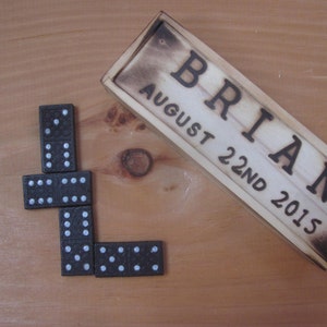 May include: A wooden box with the name "BRIAN" and the date "AUGUST 22ND 2015" engraved on it. A set of black dominoes are arranged in a L shape on a wooden surface.
