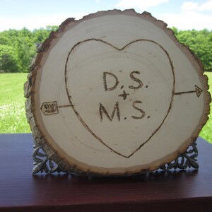 Może przedstawiać: Drewniany znak w kształcie serca z wypalonymi na drewnie inicjałami "D.S. + M.S.". Znak stoi na drewnianym stojaku i ma strzałkę skierowaną w prawo.