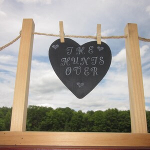 May include: A wooden sign with the text "THE HUNTS OVER" in white script, shaped like a heart, hangs from a clothesline. The sign is held up by a wooden frame. The background features a blue sky and green trees.
