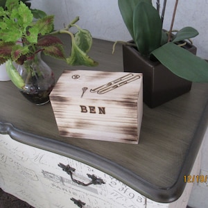 May include: A wooden box with a burnt finish and a personalised inscription that reads "BEN". The box has a design of skis and a golf club on the top.