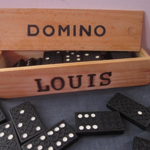 May include: A wooden box with the word "DOMINO" on the lid and "LOUIS" on the front. The box is open and contains a set of black dominoes.