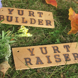 Könnte beinhalten: Zwei Holzschilder mit dem Text "Yurt Builder" und "Yurt Raiser", der in das Holz gebrannt ist. Die Schilder liegen auf einem Bett aus grünem Gras mit einigen Herbstblättern, die verstreut sind.