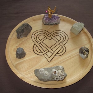 May include: A wooden tray with a Celtic knot heart design, featuring a small golden figurine and various stones, including amethyst, grey, and brown.
