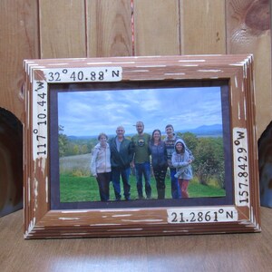 May include: A wooden picture frame containing a photograph of a family of six standing in front of a scenic view. The frame has coordinates inscribed on each side: 32° 40.88' N, 117° 10.44' W, 21.2861° N, 157.8429° W.