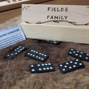 May include: A wooden box with the words "FIELDS FAMILY" burnt into the front. The box is sitting on a brown fur surface with black dominoes scattered around it. The box is open and a small piece of paper with the words "RULES FOR PLAYING DOMINOES" is visible.