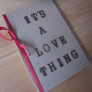 Puede incluir: Un cuaderno marrón con una cinta roja atada alrededor. El cuaderno tiene las palabras "It's a Love Thing" impresas en negro en la portada.