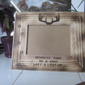 May include: A rustic wooden picture frame with a burnt wood finish. The frame has a deer antler design and the text "Memories Made On A Hunt Last A Lifetime".