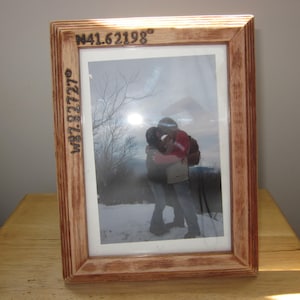 May include: A wooden picture frame with a photo of a couple embracing in a snowy landscape. The frame is engraved with the coordinates "N41.62198° W82.12727°".