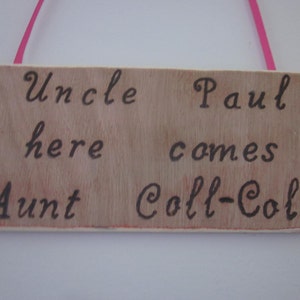 May include: A wooden sign with the text "Uncle Paul here comes Aunt Coll-Coll" written in black cursive. The sign is hanging from a pink ribbon.