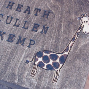 Puede incluir: Una caja de madera con los nombres "Heath Cullen Kemp" grabados en la parte delantera. Una jirafa marrón y negra está quemada en la madera.