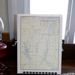 May include: A vintage map of Traverse City, Michigan, focusing on Silver Lake. The map is printed on white paper and features blue lines for roads and waterways. The map is framed and displayed on a wooden surface.