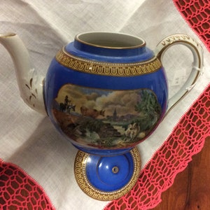 May include: A blue and white porcelain teapot with a landscape scene. The teapot has a round body, a curved spout, and a handle. Gold trim accents the rim and base. The teapot is displayed on a white cloth with a red lace trim.