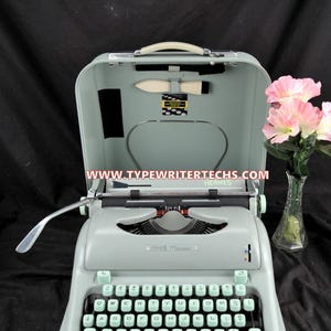 May include: A light sage green Hermes Media 3 portable typewriter with a matching hard-shell carrying case lid. The typewriter features a QWERTY keyboard with light green keys, a metal carriage return lever, and a cleaning brush inside the lid.