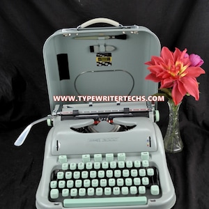 May include: A light seafoam green Hermes 3000 typewriter with a matching carrying case lid featuring a cleaning brush and the text WWW.TYPEWRITERTECHS.COM. The typewriter has a QWERTY keyboard with light green keys and a silver metal carriage.