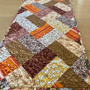 May include: A handmade, quilted table runner with a patchwork design. The runner features a variety of fabrics in shades of brown, orange, yellow, and cream, arranged in a diagonal pattern. The edges are finished with a floral print.