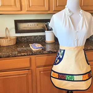 May include: A beige apron with a blue and white floral print and a colorful patchwork border. The apron is on a white mannequin with a white blouse. The background is a kitchen with wooden cabinets and a granite countertop.