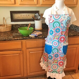 May include: A colorful kitchen apron with a blue pocket and white ruffle trim. The apron features a pattern of pink, blue, green, and yellow circles with floral designs. The apron is hanging on a white mannequin in a kitchen setting.