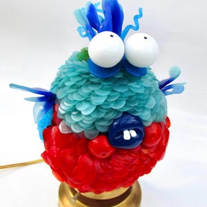 Puede incluir: Una lámpara caprichosa con forma de pez de dibujos animados. La lámpara presenta un cuerpo azul y turquesa, ojos blancos grandes y una base roja. El pez tiene una boca azul y está montado sobre una base dorada. Un artículo decorativo único.
