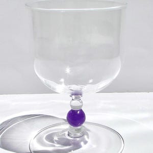May include: A clear glass goblet with a wide bowl and a purple ball accent on the stem. The goblet sits on a circular base. The glass casts a shadow on the white surface.