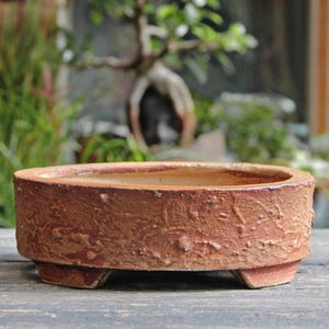 May include: A low, round, reddish-brown ceramic bonsai pot with a textured exterior and three small feet. The pot has a wide, open top and is designed for planting small trees or plants. The background shows a blurred bonsai tree.