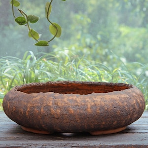May include: A low, round, brown ceramic bowl with a rough, textured exterior and a wide, open top. The vessel features a dark, weathered finish with visible cracks and an uneven, organic rim, resting on a flat base on a wooden surface.