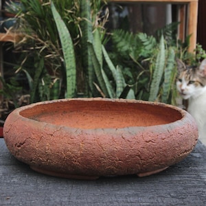 May include: A shallow, round terracotta bowl with a thick, textured, and cracked exterior surface. The interior of the bowl is smooth and features a consistent reddish-brown clay color, while the rim is rounded and substantial in its overall form.