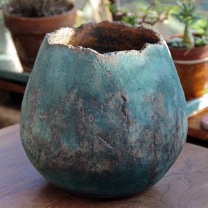 May include: A teal-colored, handcrafted ceramic vase with a textured surface and a unique, uneven rim. The vase has a rounded shape and a dark interior, suggesting it could be used for decorative purposes or as a planter. The vase is sitting on a wooden surface.