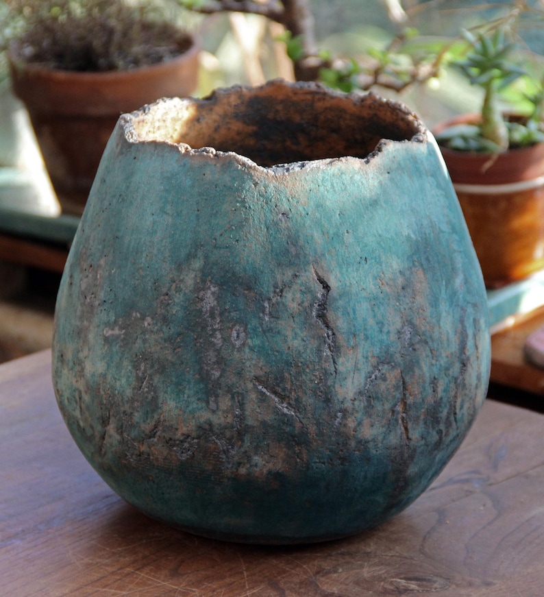 May include: A teal-colored, handcrafted ceramic vase with a textured surface and a unique, uneven rim. The vase has a rounded shape and a dark interior, suggesting it could be used for decorative purposes or as a planter. The vase is sitting on a wooden surface.