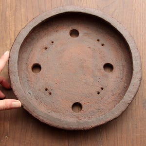 May include: A terracotta saucer with a round shape and multiple drainage holes. The saucer is a reddish-brown color and has a rough texture.