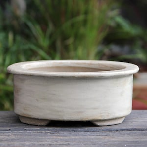 Könnte beinhalten: Ein cremefarbener, oval geformter Bonsai-Topf mit abgerundetem Rand und drei kleinen Füßen. Der Topf hat eine strukturierte Oberfläche und eine neutrale Farbpalette, die für verschiedene Pflanzenarten geeignet ist. Der Topf steht auf einer Holzoberfläche.