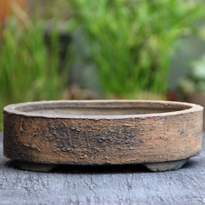 May include: A brown oval ceramic bonsai pot with a rough, textured surface. The pot has three small feet on the bottom.