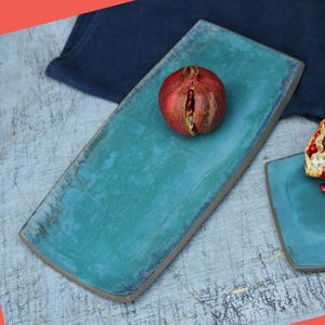 Peut inclure: Un plat de service rectangulaire en céramique turquoise avec une grenade. Le plat a une surface texturée et un bord gris. La grenade est rouge avec des graines visibles. L'arrière-plan comprend un tissu bleu et un second plat.