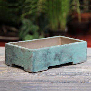 Könnte beinhalten: Ein rechteckiger, türkisfarbener Keramik-Pflanztopf mit verwittertem Aussehen. Der Pflanztopf hat einen hellbraunen Innenraum und steht auf vier kleinen Füßen. Die Außenseite hat ein gesprenkeltes, gealtertes Aussehen, was auf Witterungseinflüsse hindeutet.