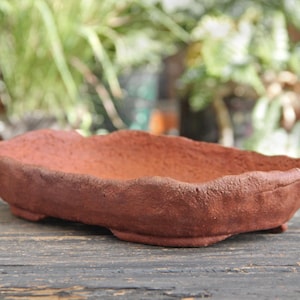 Puede incluir: Un tiesto para bonsái hecho a mano, de color terracota, con una superficie texturizada y un borde irregular. El recipiente rectangular tiene un perfil bajo y se apoya sobre pequeños pies, ideal para plantas pequeñas o árboles en miniatura.