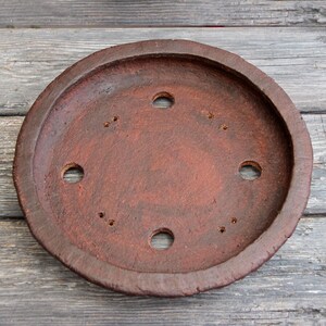 May include: A terracotta colored round saucer with six drainage holes. The saucer is made of a rough, textured material.