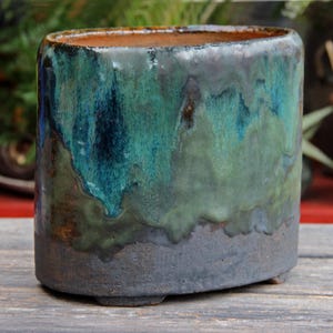 Op de afbeelding: Een grote ovale keramische pot met een donkergroene en blauwe glazuur. De pot heeft een ruw, getextureerd oppervlak en een donkergrijze basis.