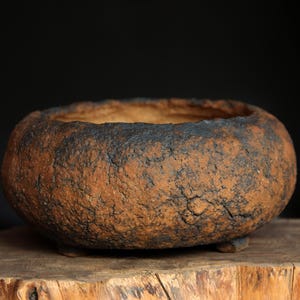 Può includere: Un vaso per bonsai rotondo in ceramica fatto a mano. Il vaso ha una superficie ruvida e strutturata con una combinazione di colori marrone scuro e nero. Il vaso per bonsai poggia su una superficie di legno e ha tre piedini.