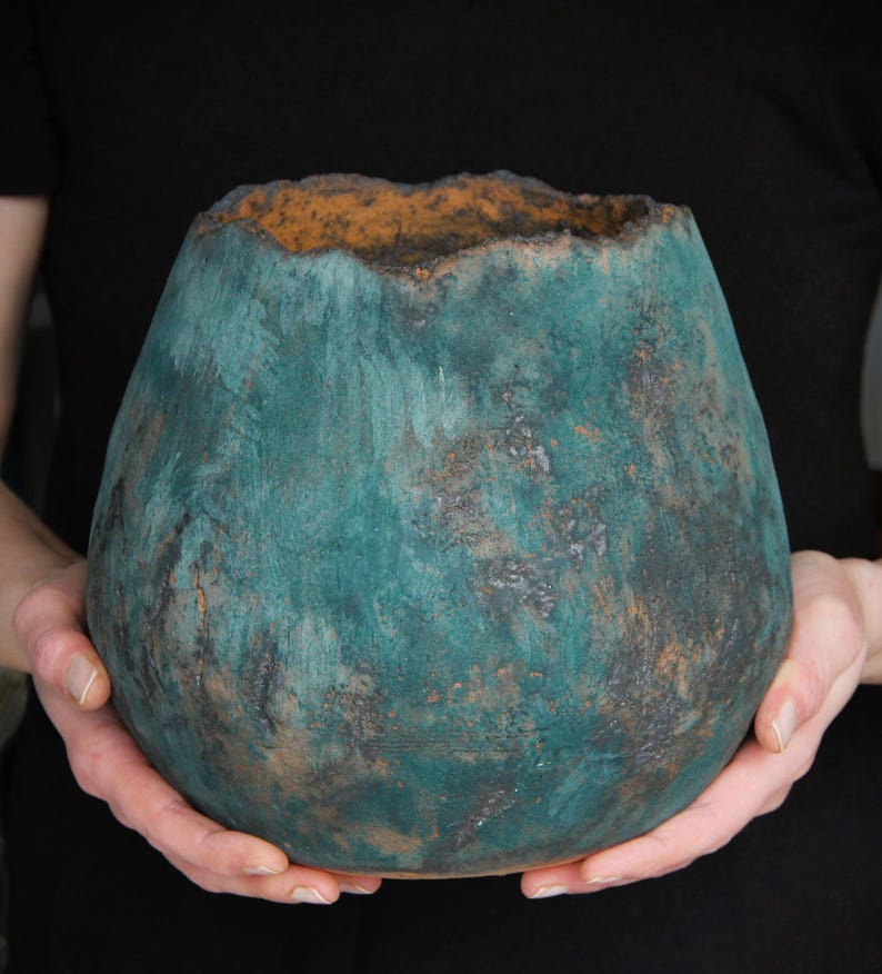 May include: A handcrafted, teal-colored ceramic vase with a textured surface and a unique, uneven rim. The vase has a rounded, bulbous shape and is held by two hands against a black background. The interior rim is a warm, earthy orange.